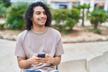 Sticker - Young latin man using smartphone sitting on bench at park