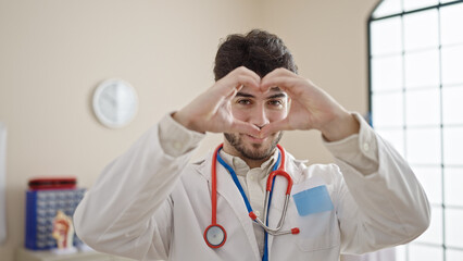 Wall Mural - Young hispanic man doctor smiling doing heart gesture with hands at clinic