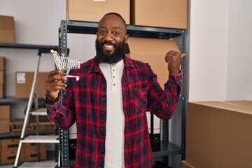 Poster - African american man working at small business ecommerce holding cart pointing thumb up to the side smiling happy with open mouth