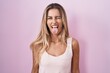 Young blonde woman standing over pink background sticking tongue out happy with funny expression. emotion concept.
