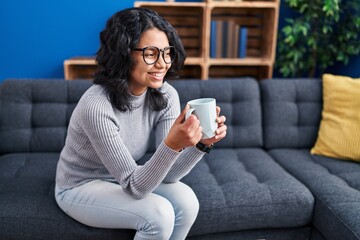 Sticker - Young latin woman drinking coffee sitting on sofa at home