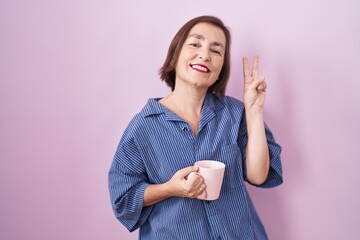 Poster - Middle age hispanic woman drinking a cup coffee smiling looking to the camera showing fingers doing victory sign. number two.