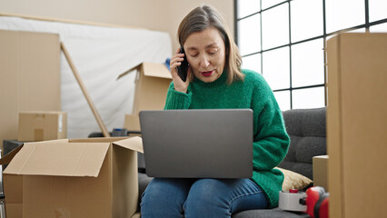 Sticker - Mature hispanic woman with grey hair talking on smartphone using laptop at new home