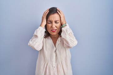 Poster - Middle age hispanic woman standing over blue background suffering from headache desperate and stressed because pain and migraine. hands on head.