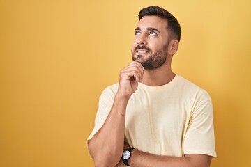 Wall Mural - Handsome hispanic man standing over yellow background with hand on chin thinking about question, pensive expression. smiling and thoughtful face. doubt concept.