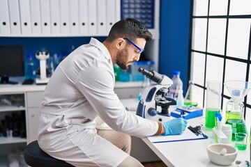 Sticker - Young hispanic man wearing scientist uniform using microscope write on document at laboratory