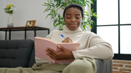 Poster - African american woman writing on notebook sitting on sofa at home