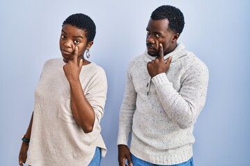 Sticker - Young african american couple standing over blue background together pointing to the eye watching you gesture, suspicious expression