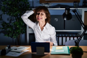 Wall Mural - Middle age woman working at the office at night smiling confident touching hair with hand up gesture, posing attractive and fashionable