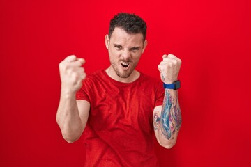 Sticker - Young hispanic man standing over red background angry and mad raising fists frustrated and furious while shouting with anger. rage and aggressive concept.