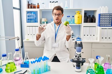 Poster - Young hispanic man working at scientist laboratory holding apple in shock face, looking skeptical and sarcastic, surprised with open mouth