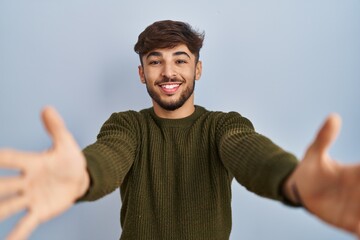 Sticker - Arab man with beard standing over blue background looking at the camera smiling with open arms for hug. cheerful expression embracing happiness.