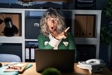 Sticker - Middle age woman working at night using computer laptop surprised pointing with finger to the side, open mouth amazed expression.