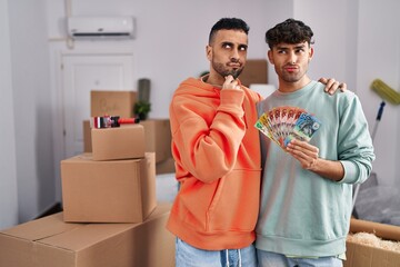 Wall Mural - Young hispanic gay couple moving to a new home holding banknotes serious face thinking about question with hand on chin, thoughtful about confusing idea