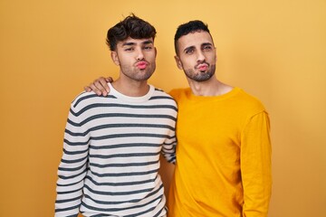 Wall Mural - Young hispanic gay couple standing over yellow background looking at the camera blowing a kiss on air being lovely and sexy. love expression.