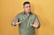 Hispanic young man standing over yellow background moving away hands palms showing refusal and denial with afraid and disgusting expression. stop and forbidden.