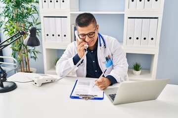 Canvas Print - Young latin man doctor talking on telephone writing on document at clinic