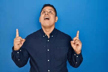 Canvas Print - Hispanic young man standing over blue background amazed and surprised looking up and pointing with fingers and raised arms.