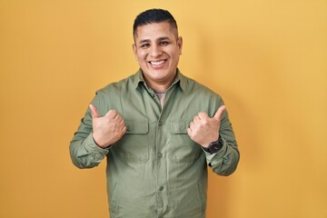 Canvas Print - Hispanic young man standing over yellow background success sign doing positive gesture with hand, thumbs up smiling and happy. cheerful expression and winner gesture.