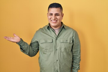 Canvas Print - Hispanic young man standing over yellow background smiling cheerful presenting and pointing with palm of hand looking at the camera.