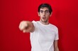 Young hispanic man standing over red background pointing with finger surprised ahead, open mouth amazed expression, something on the front