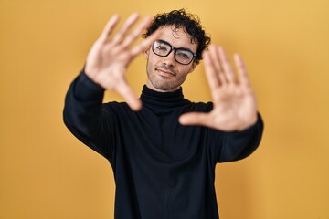 Wall Mural - Hispanic man standing over yellow background doing frame using hands palms and fingers, camera perspective