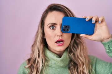 Sticker - Young caucasian woman covering eye with smartphone in shock face, looking skeptical and sarcastic, surprised with open mouth