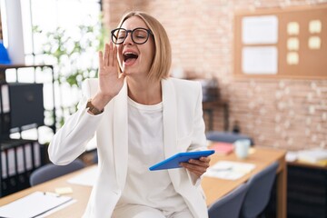 Canvas Print - Young caucasian woman working at the office wearing glasses shouting and screaming loud to side with hand on mouth. communication concept.