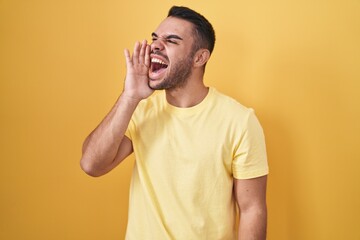 Canvas Print - Young hispanic man standing over yellow background shouting and screaming loud to side with hand on mouth. communication concept.