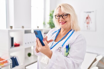Wall Mural - Middle age blonde woman wearing doctor uniform using smartphone at clinic