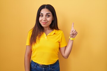 Wall Mural - Young arab woman standing over yellow background showing and pointing up with finger number one while smiling confident and happy.