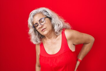 Poster - Middle age woman with grey hair standing over red background suffering of backache, touching back with hand, muscular pain