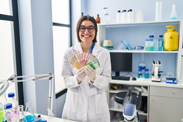 Canvas Print - Young hispanic woman working at scientist laboratory holding money banknotes celebrating crazy and amazed for success with open eyes screaming excited.