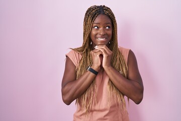 Poster - African american woman with braided hair standing over pink background laughing nervous and excited with hands on chin looking to the side