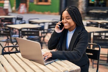 Poster - African american woman talking on smartphone using laptop sitting on table at coffee shop terrace