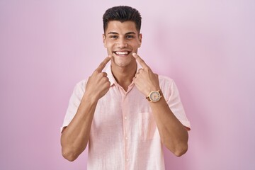 Wall Mural - Young hispanic man standing over pink background smiling with open mouth, fingers pointing and forcing cheerful smile