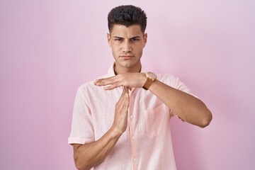 Wall Mural - Young hispanic man standing over pink background doing time out gesture with hands, frustrated and serious face