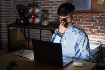 Canvas Print - Young hispanic man with beard working at the office at night smelling something stinky and disgusting, intolerable smell, holding breath with fingers on nose. bad smell