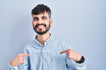 Sticker - Young hispanic man with beard standing over blue background looking confident with smile on face, pointing oneself with fingers proud and happy.