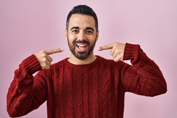 Wall Mural - Young hispanic man with beard wearing casual sweater over pink background smiling cheerful showing and pointing with fingers teeth and mouth. dental health concept.