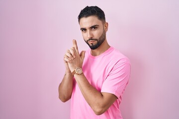 Poster - Hispanic young man standing over pink background holding symbolic gun with hand gesture, playing killing shooting weapons, angry face