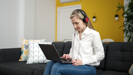 Sticker - Young blonde woman using laptop and headphones sitting on sofa at home