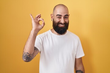 Canvas Print - Young hispanic man with beard and tattoos standing over yellow background smiling positive doing ok sign with hand and fingers. successful expression.