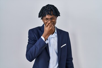 Poster - Young african man with dreadlocks wearing business jacket over white background smelling something stinky and disgusting, intolerable smell, holding breath with fingers on nose. bad smell