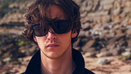 Portrait of an young man with brown hair blown on wind against rock on summer sunny day. Attractive young man looks at the camera on the nature. Handsome guy in black sunglasses poses outdoors.