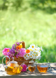 Fototapeta Nowy Jork - flowers in basket, glass teapot and cups with herbal tea on table, natural abstract background. summer season. Beautiful rustic composition. relax time. useful calming tea. Tea party in garden