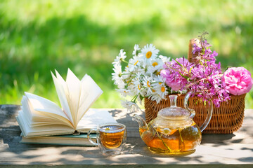 Sticker - glass teapot and cup with herbal tea, flowers in basket, book close up on table, natural abstract background. Beautiful rustic composition. relax time. useful calming tea. Tea party in garden