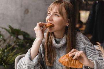 Young blonde woman with bang eating croissants at a cafe. Girl bite piece of croissant look joyful at restaurant. Cheat meal day concept. Woman is preparing with appetite to eat croissant.