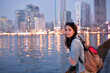 © Ekaterina - A tourist girl with a backpack on her shoulders enjoys a view of the modern skyscrapers of the Sharjah marina at night