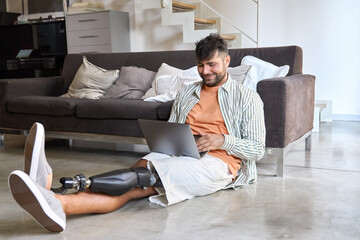 Happy young man with disability leg prosthesis sitting on floor at home using laptop computer working or elearning, browsing web, searching online. People with amputation disabilities everyday life.